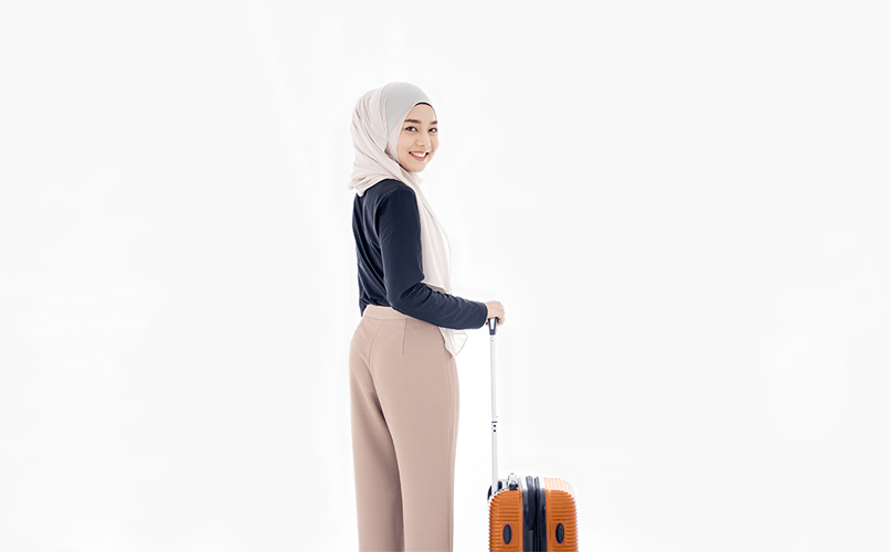 A woman wearing a hijab, dark long-sleeve shirt, and light-colored trousers is smiling while standing with an orange wheeled suitcase. She is facing sideways but looking towards the camera against a plain white background, ready to get a job abroad.