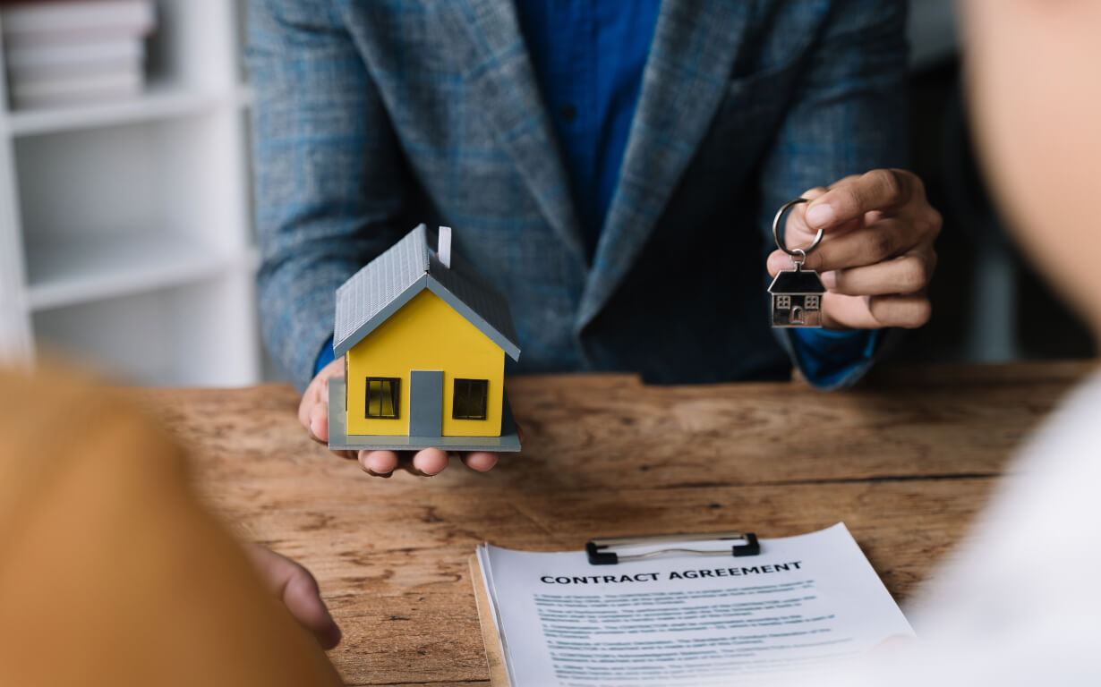 Une personne en costume tient une petite maquette de maison jaune et des clés, symbolisant l’achat d’une propriété à l’étranger, tandis qu’un autre individu est assis en face d’une table en bois avec un contrat visible, préparant le terrain pour une transaction immobilière internationale.